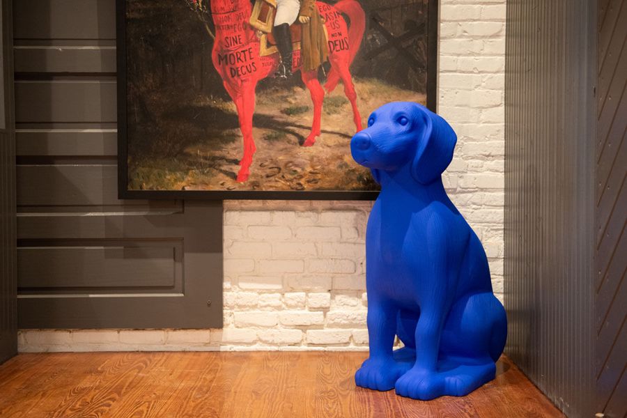 Cobalt blue dog sculpture seated beneath a painting of a rider on a red horse against whitewashed brick