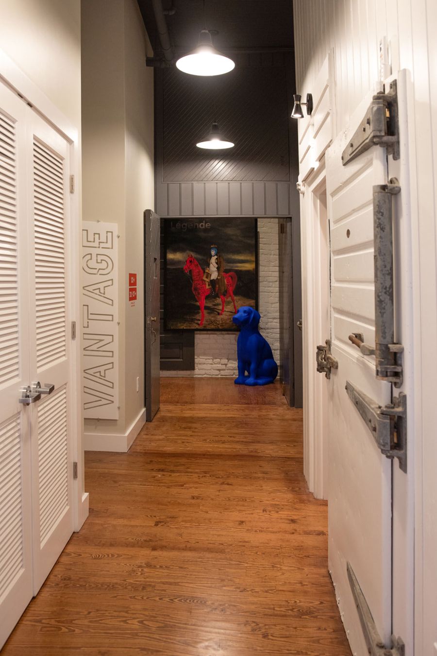Guest hallway with heart-pine floors, a VANTAGE wall sign and a blue dog sculpture beneath a painting at the far end