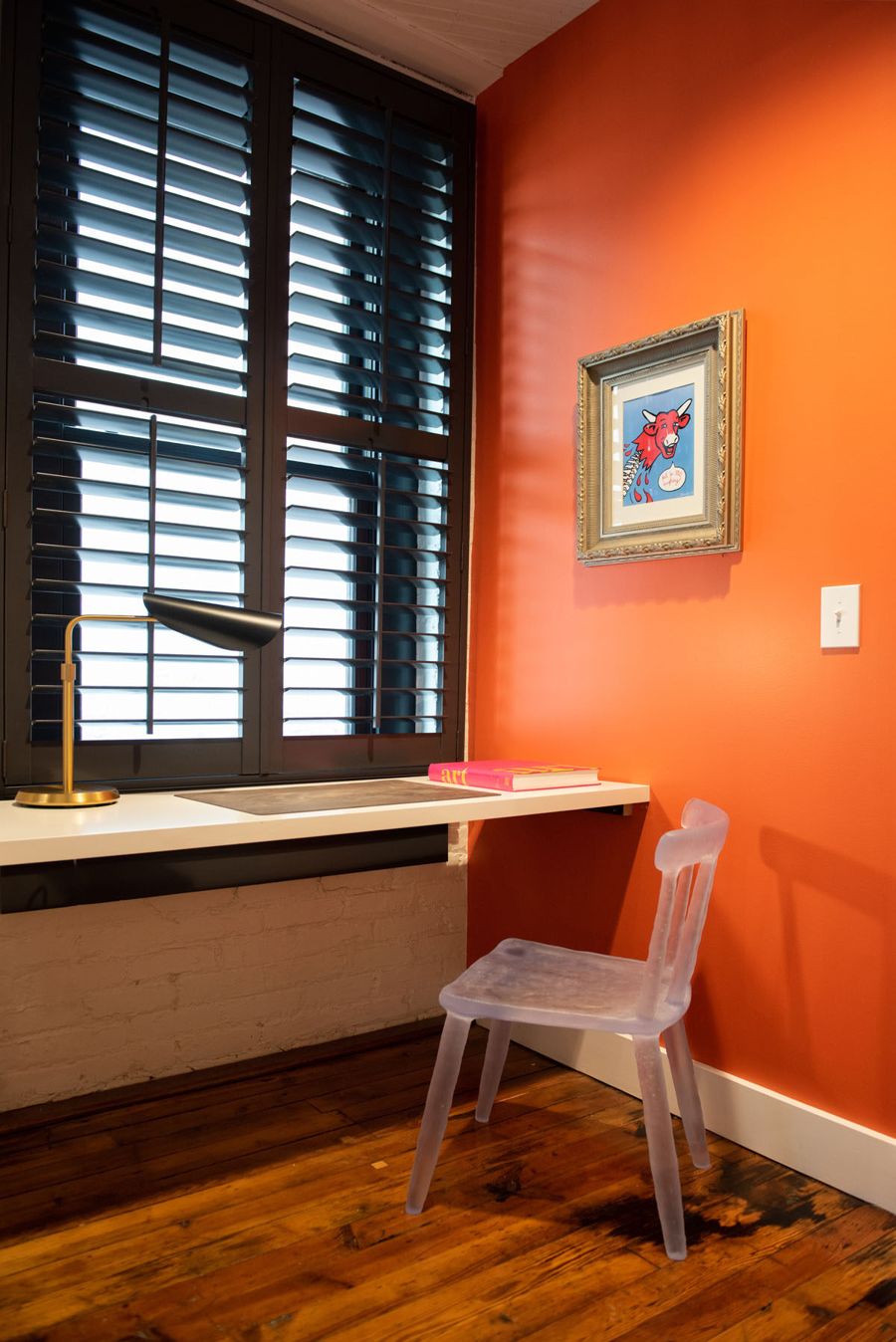 Dedicated workstation with a clear acrylic chair beside an orange wall and shuttered window
