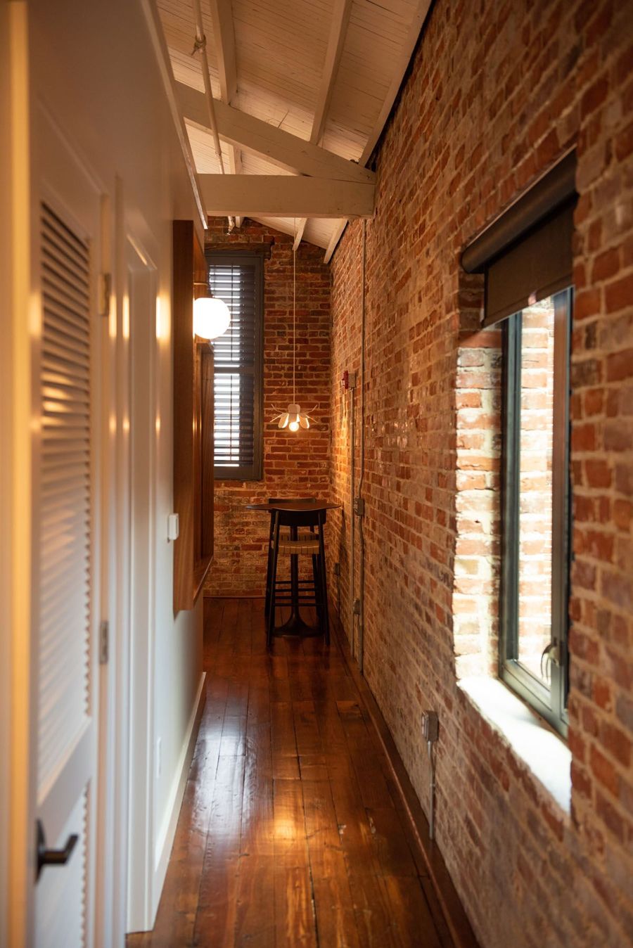 Hallway with original heart pine floors and an exposed brick wall leading to a high-top table by a shuttered window