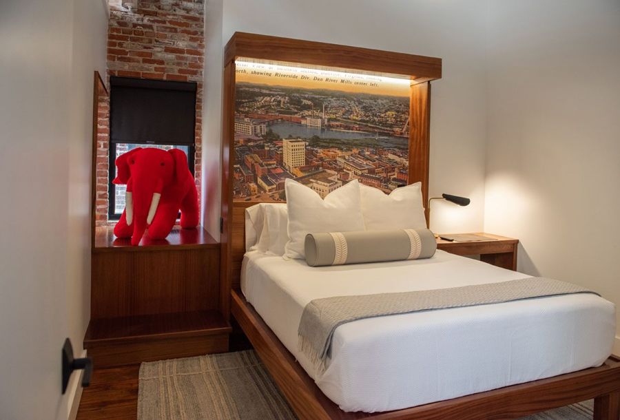 Bedroom with a backlit sapele headboard framing a vintage aerial postcard of Danville and the Dan River Mills, beside a red elephant on the window bench