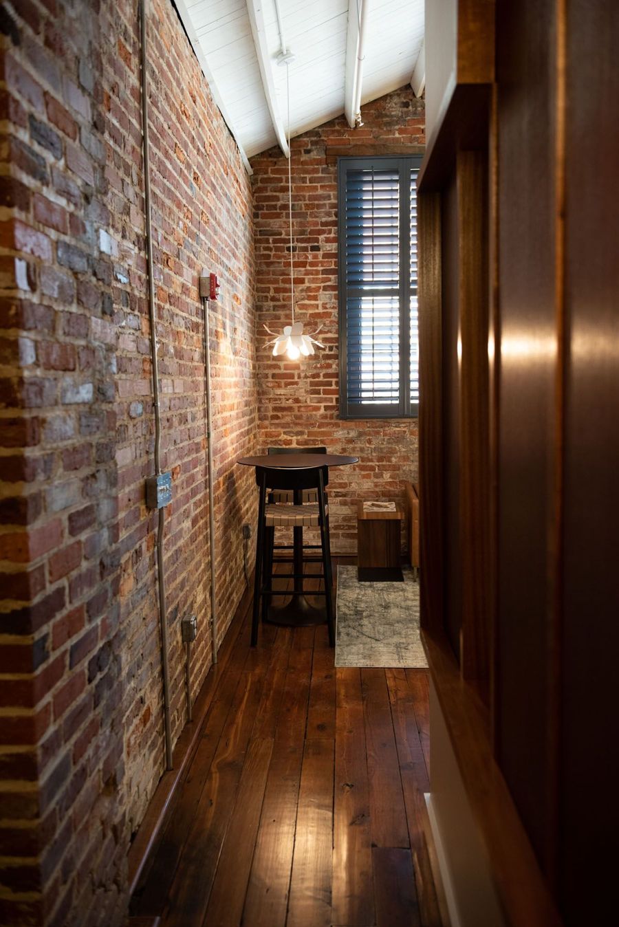 Long exposed brick wall leading past a sapele wood partition to a bistro table beside a shuttered window